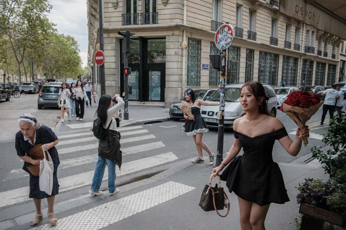 A woman in a black off-shoulder dress holding a bouquet of red roses and a handbag, standing on a city sidewalk near a crosswalk, with other pedestrians walking in the background.