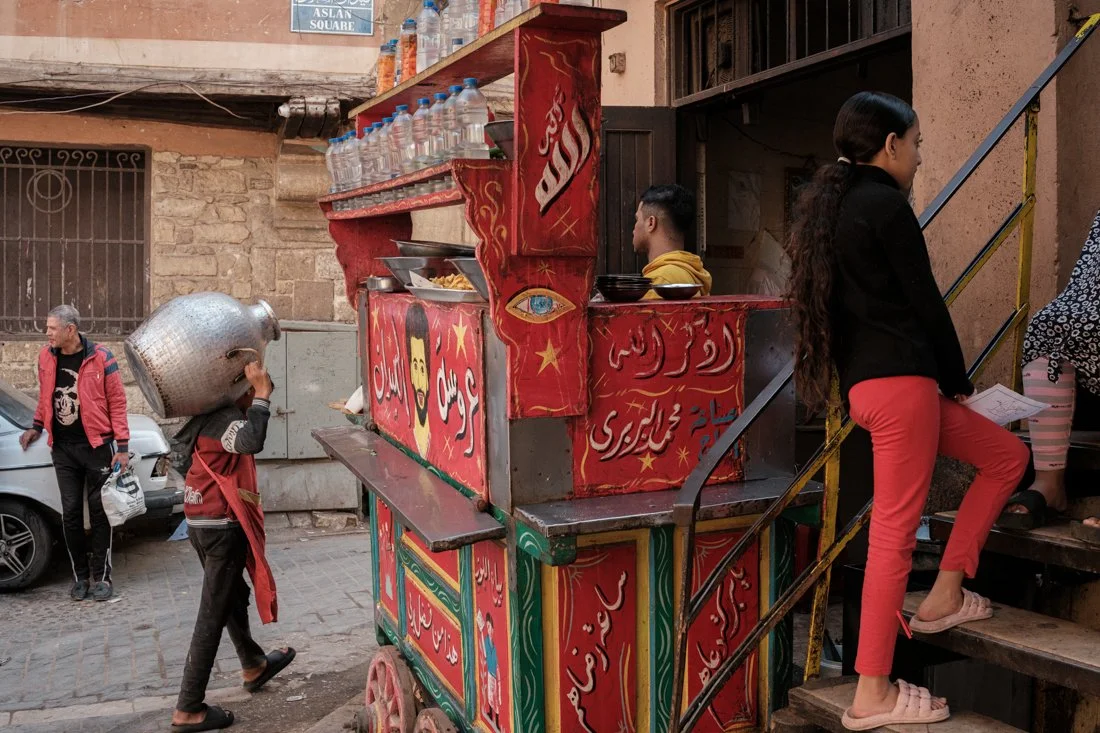 A colorful street food cart with red and green decorations, serving food, and a woman standing on stairs to buy or pick up food.