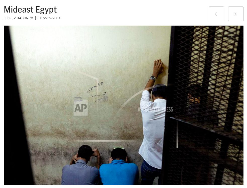 Three men sitting on the floor with one man standing in front of a worn wall, writing or scribbling on it.