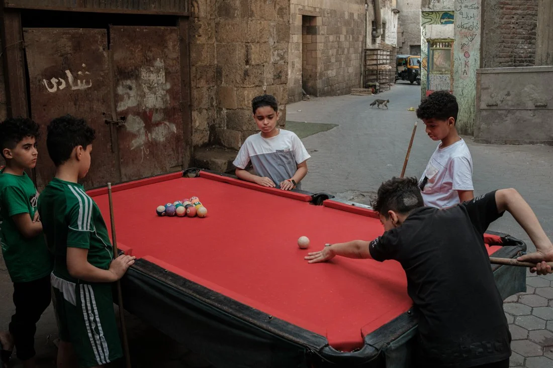 Five boys playing pool on a red pool table in an alleyway, with a small cat in the background.