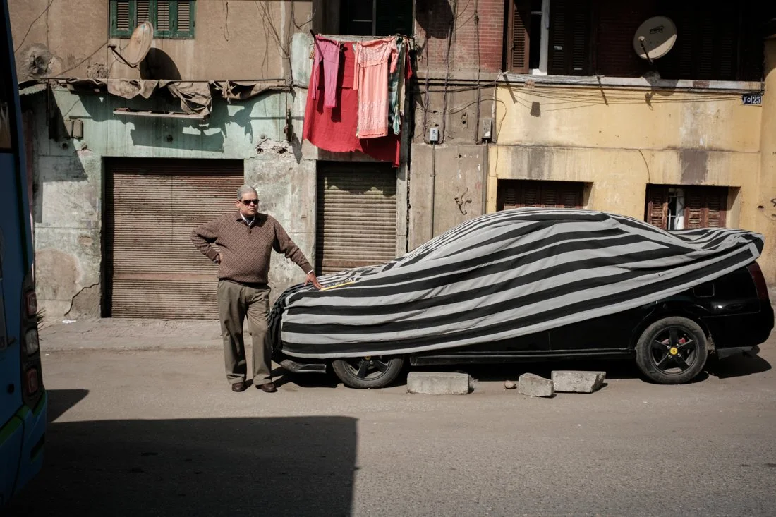 A man standing on a street next to a black car covered with a striped tarp, with old buildings and laundry hanging above in the background.