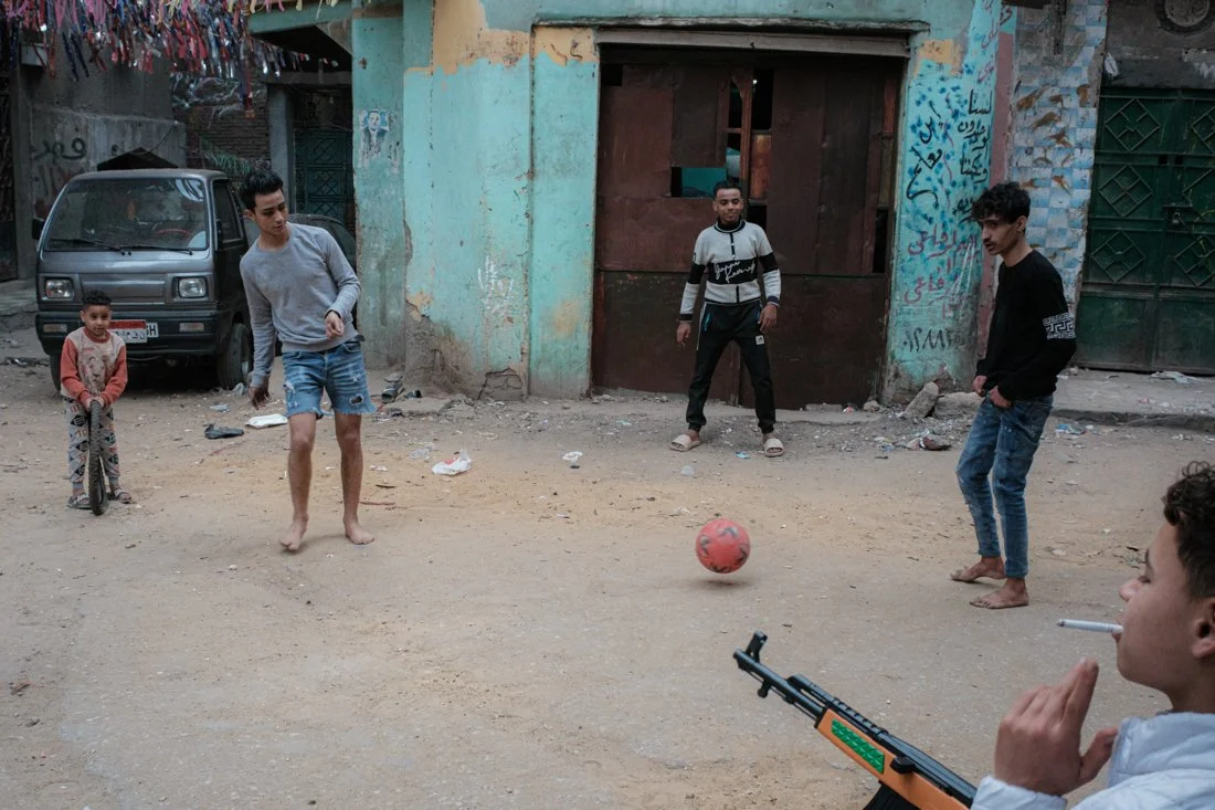 Young boys playing soccer on a dirt street in an urban neighborhood with old buildings and a parked van.