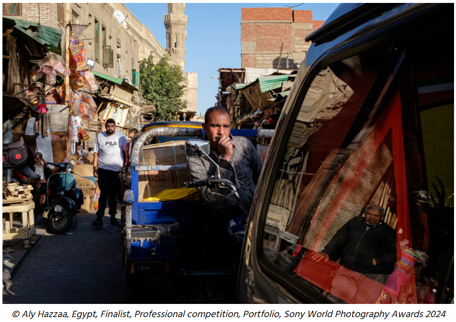 Street scene in Egypt with people walking and shopping, showing marketplace stalls and a historic tower in the background.