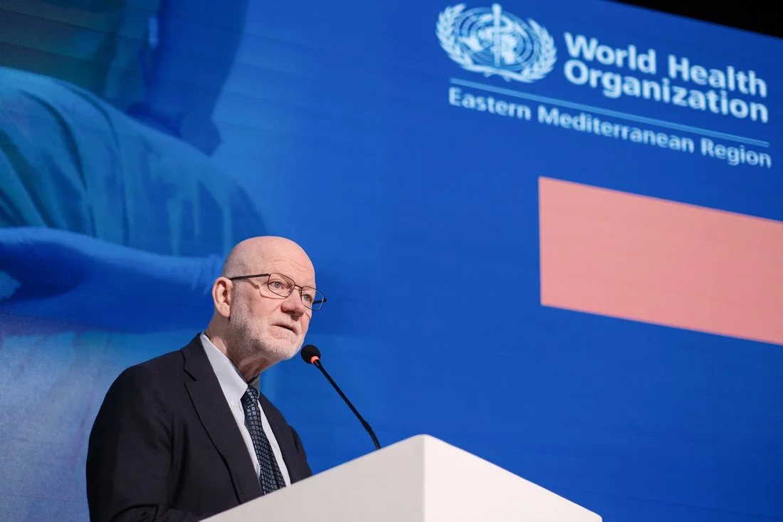 A man with glasses and a beard speaking at a podium during a World Health Organization event, with the organization's logo and a blue background behind him.