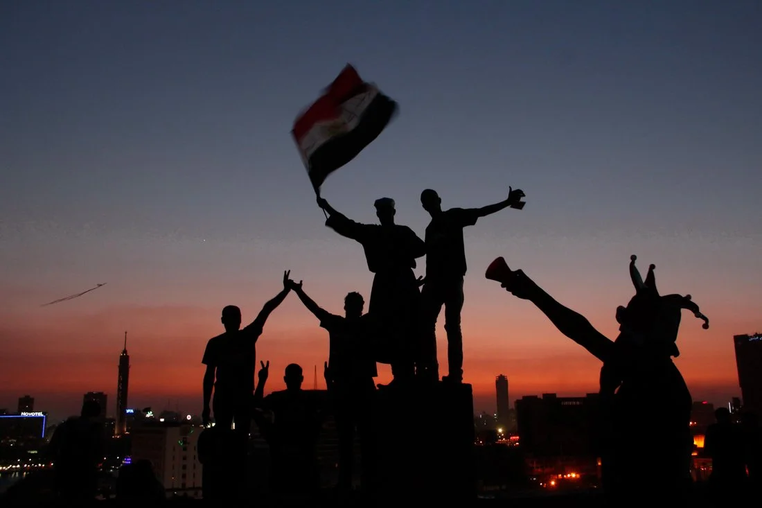 Silhouetted group of people celebrating with a flag and gestures against a sunset cityscape.