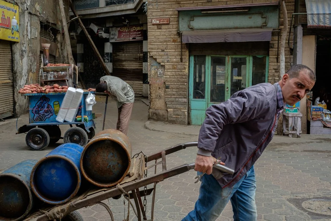 A man pushes a cart with empty rusty barrels on a cobblestone street, while another man is bending over a street vendor stand selling sweet potato, in a neighborhood with old buildings.