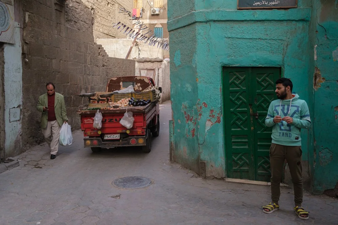 A small street scene with two men, one walking and one standing, near a red utility vehicle loaded with various goods, in front of a weathered teal-colored building with a green door.