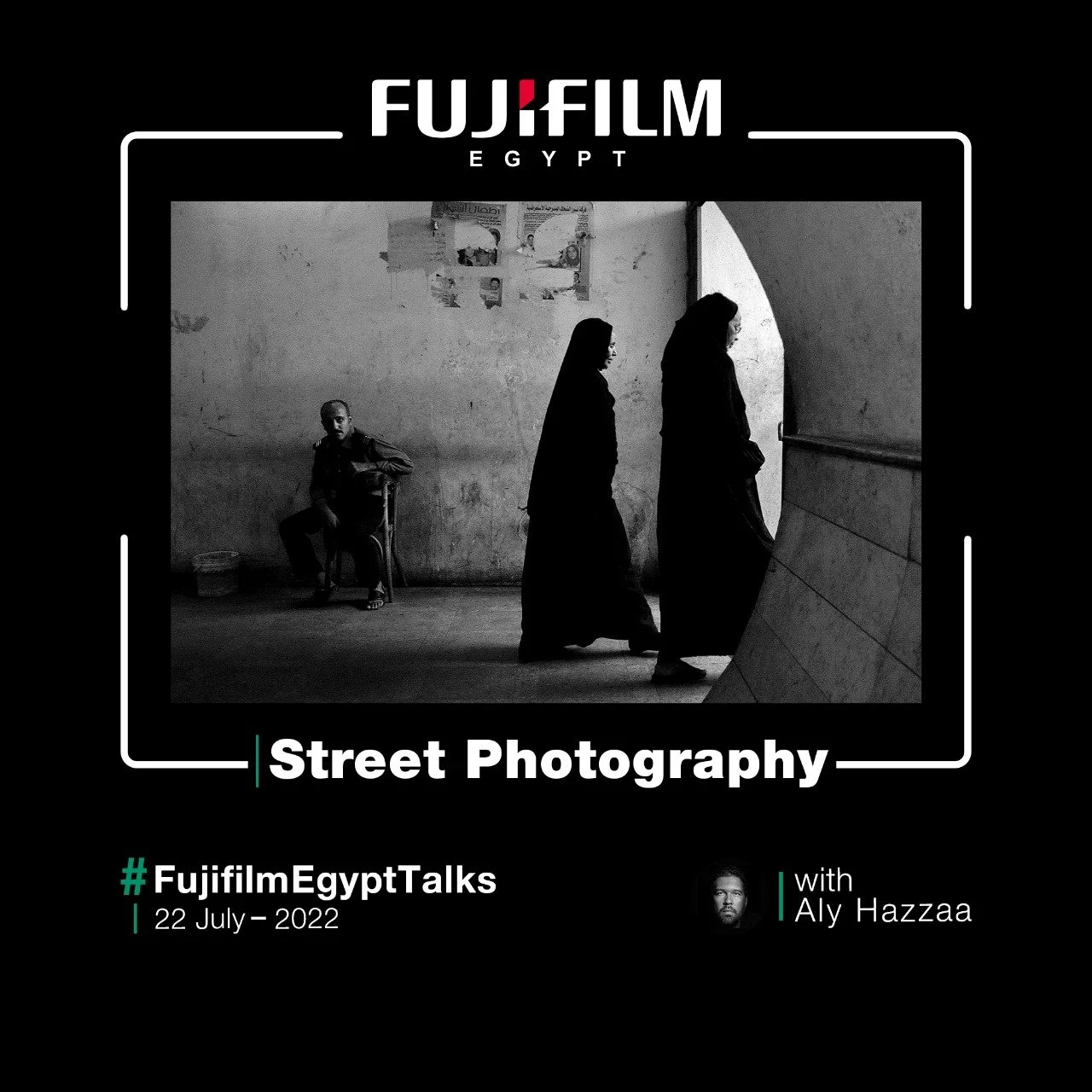 Black and white street photograph showing two women walking past a man sitting on a chair against a wall with posters in Egypt.