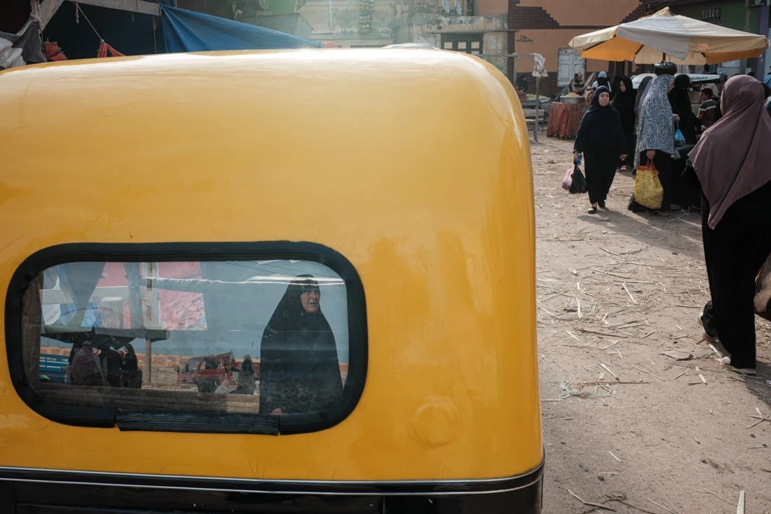 Reflected image of a person wearing a headscarf seen in the window of a yellow vehicle at an outdoor market with many people, umbrellas, and stalls in the background.