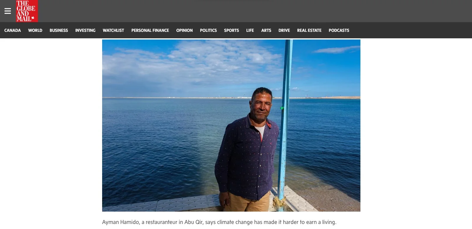 A man standing by the sea under a blue sky with clouds, holding a colorful pole, with a shoreline in the background.