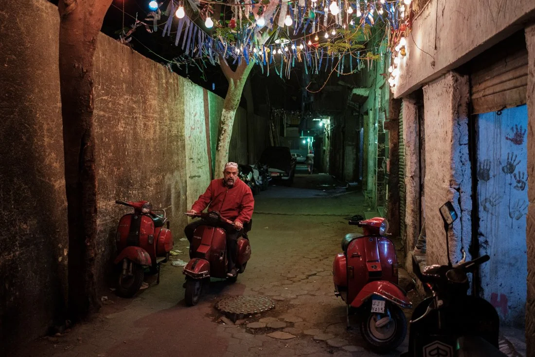 A man riding a red scooter through a narrow alleyway decorated with hanging string lights and streamers, with multiple scooters parked along the sides and painted handprints on the wall.