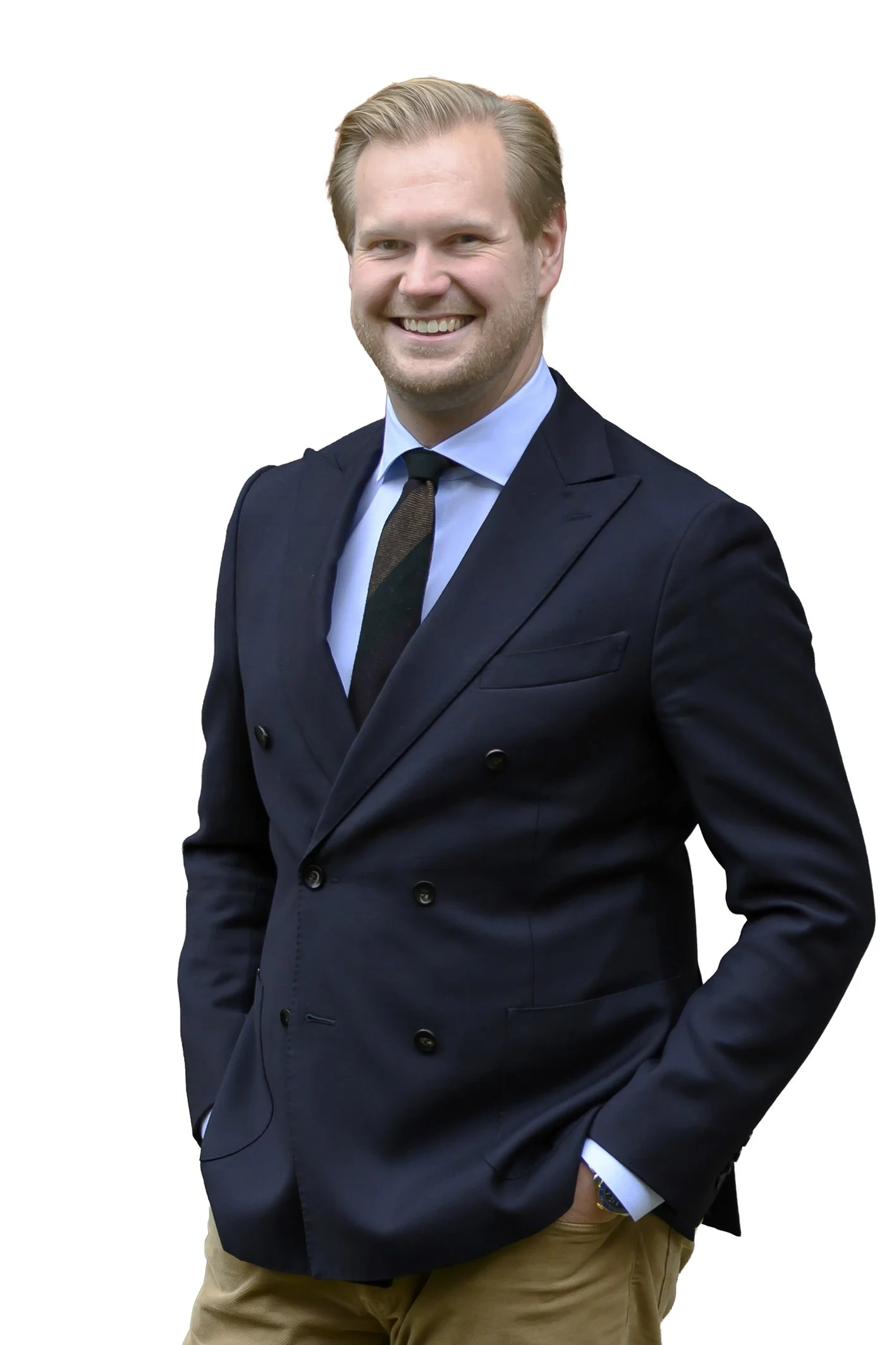 En man i kostym som ler, står med handen i fickan, bär beige byxor och en mörkblå kavaj.