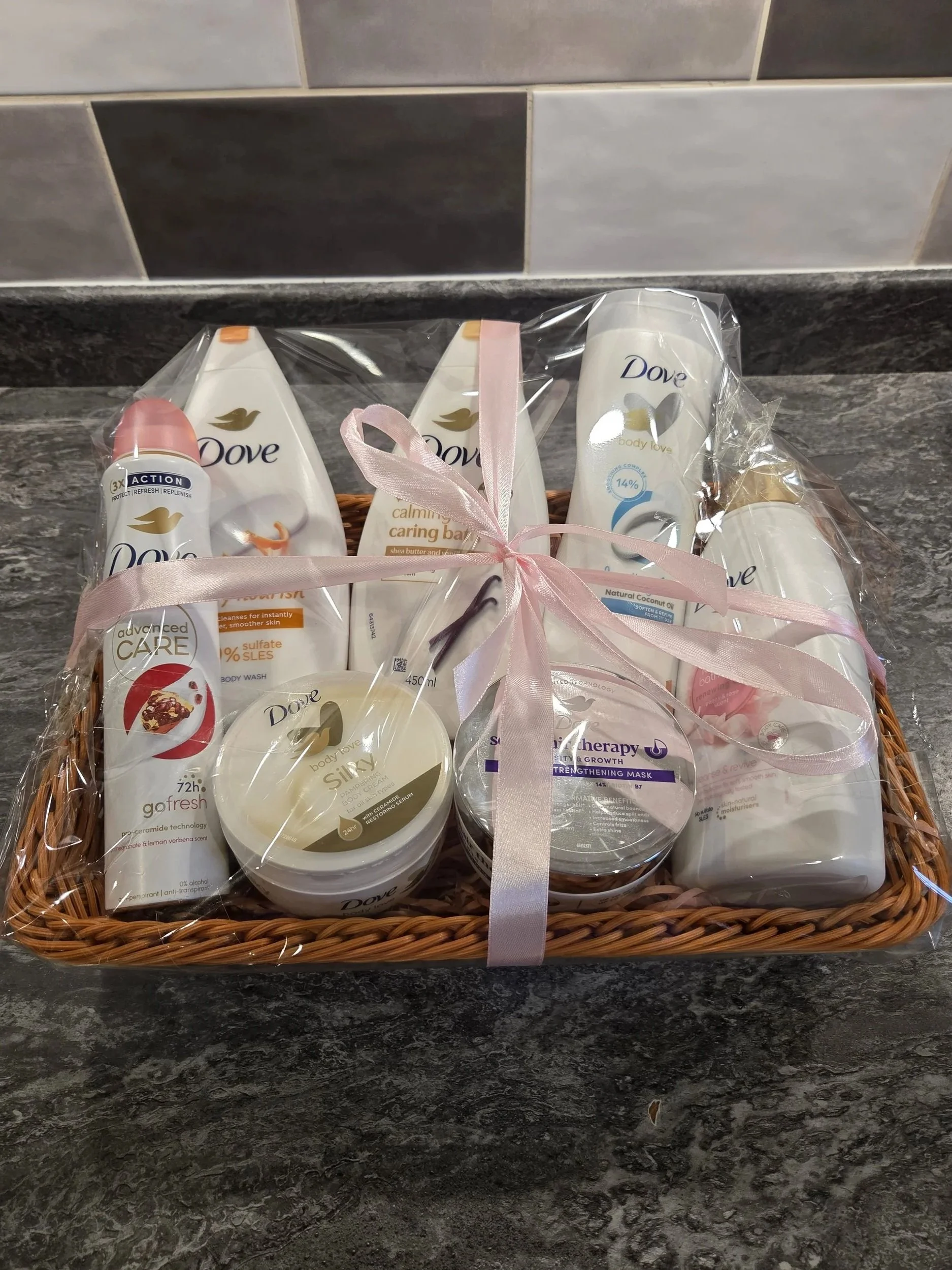 A gift basket containing various Dove skincare products wrapped in clear cellophane and tied with a pink ribbon, placed on a black marble countertop.