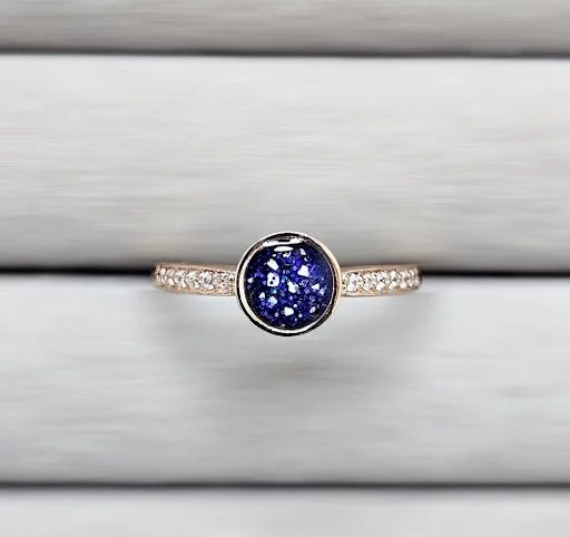 A silver ring with a dark blue glittery oval stone and small surrounding diamonds, resting on a wooden surface.