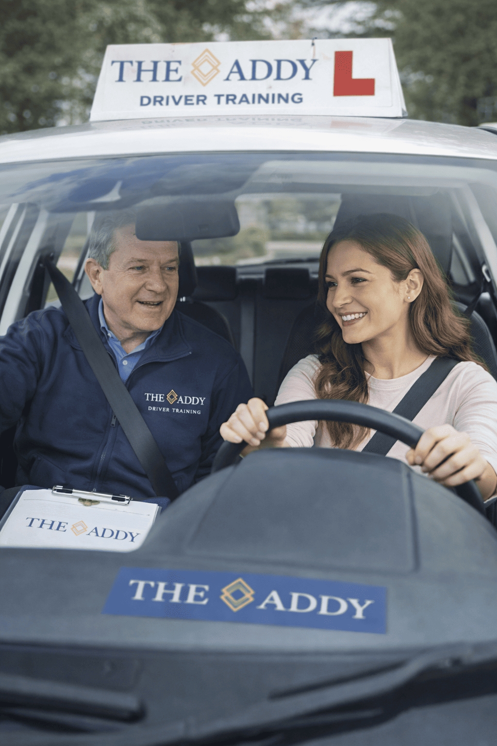 Driving instructor and student smiling inside a car during driver training, with 'THE ADDY' logo on the car and instructor's jacket.