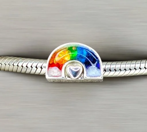 Close-up of a silver bracelet with a rainbow-colored charm in the shape of a semi-circle and a heart, resting on a light surface.