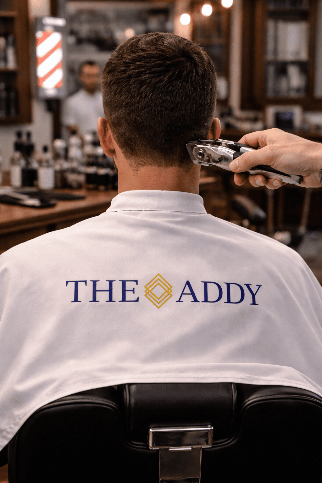 A man getting a haircut in a barbershop, with 'The Addy' embroidered on the back of his white barber cape.