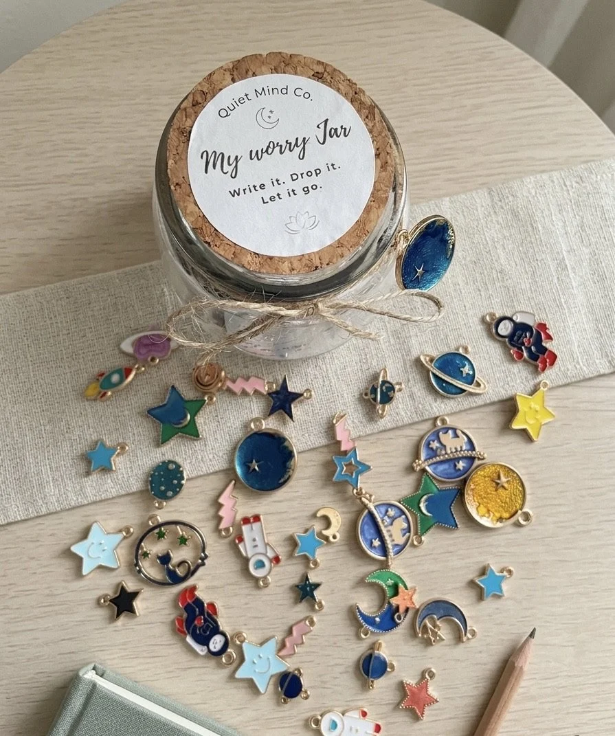 A glass jar labeled 'My worry Jar' with a cork lid, surrounded by colorful space-themed charms on a table.