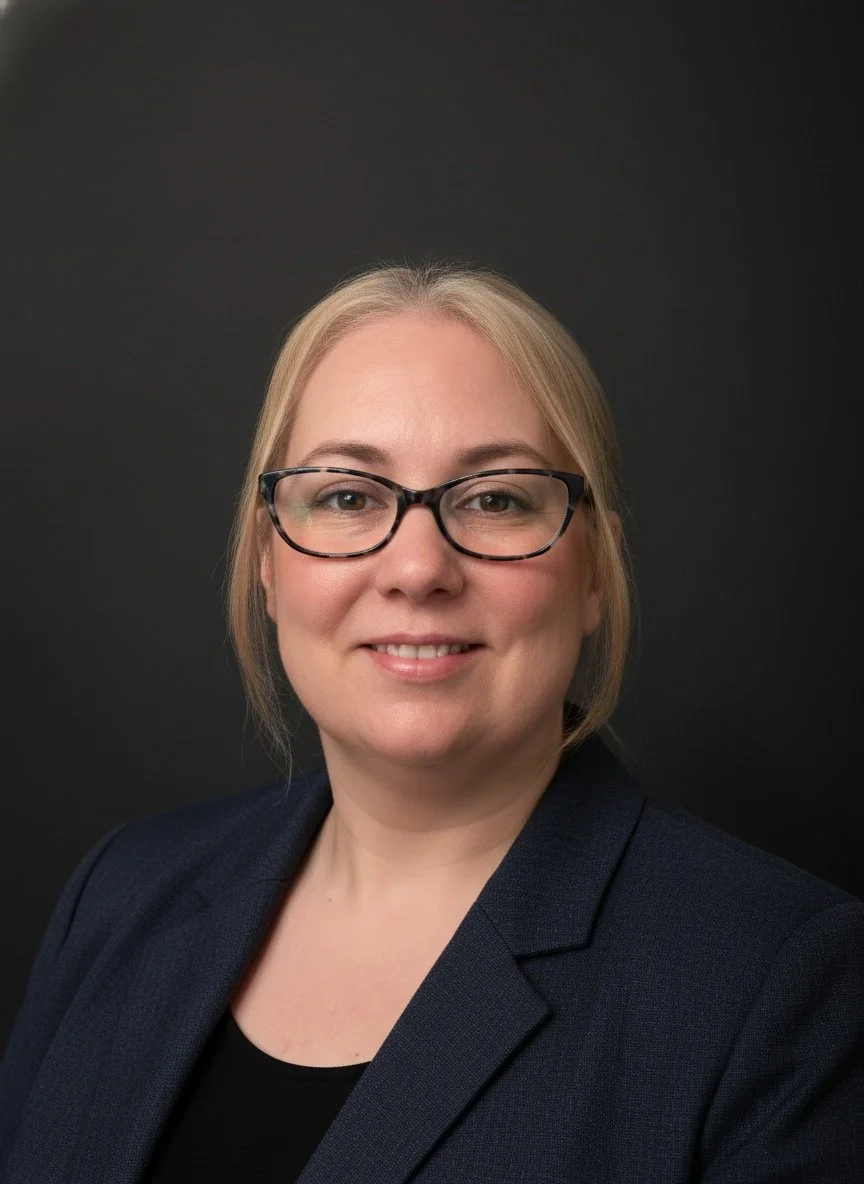 A person with glasses and blonde hair, wearing a dark blazer and smiling softly against a dark background, conveys a professional and approachable tone.