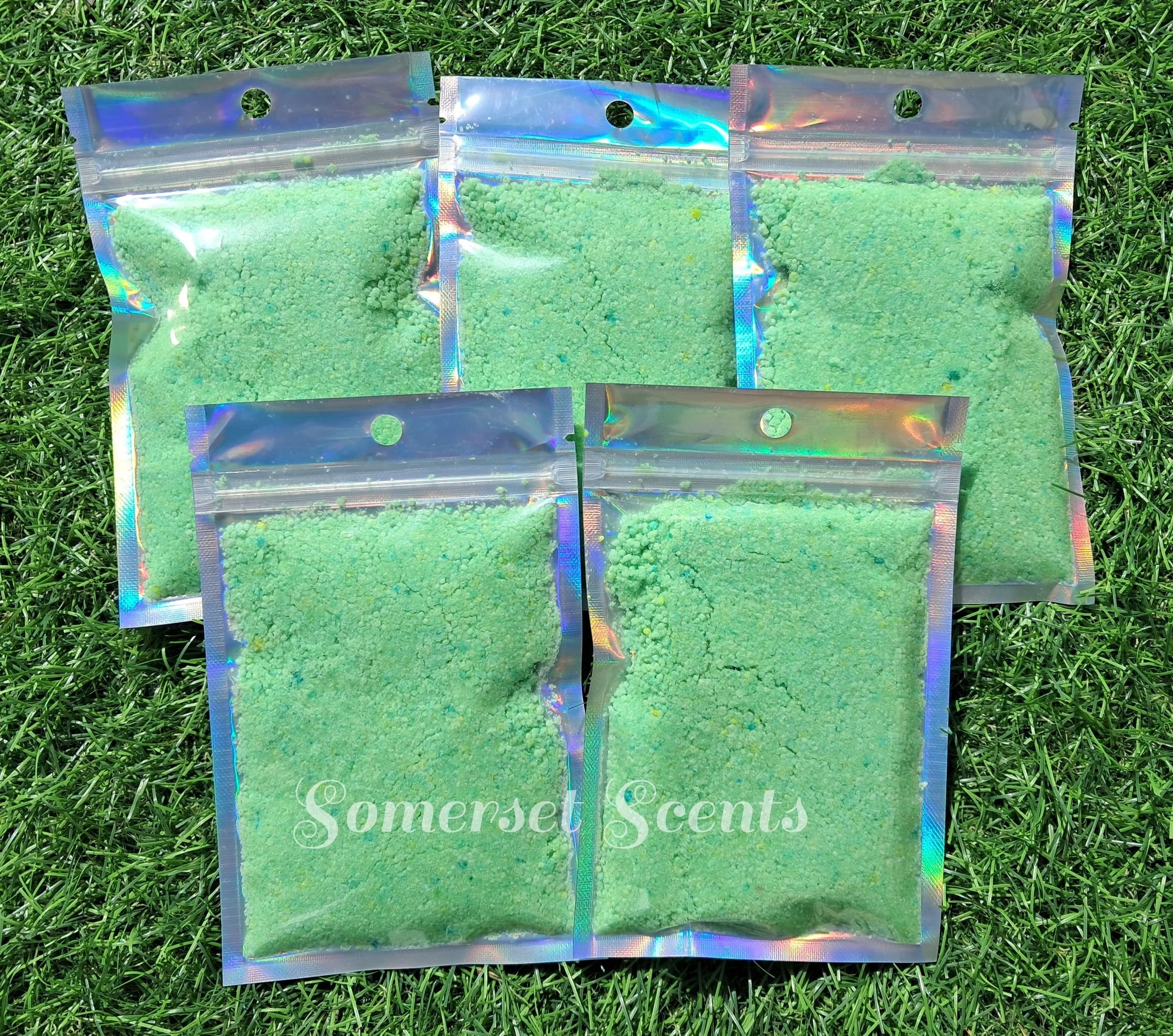 Three holographic resealable bags containing green scented bath salts labeled 'Somerset Scents', placed on green grass.