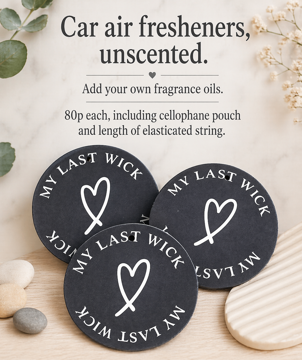 A display of three black round car air fresheners with the phrase "My Last Wick" and a heart symbol in white, along with instructions for adding fragrance oils, placed on a light wooden surface with small stones and a white striped ceramic dish nearby, and a sign above them explaining they are unscented and can be customized with fragrance oils.
