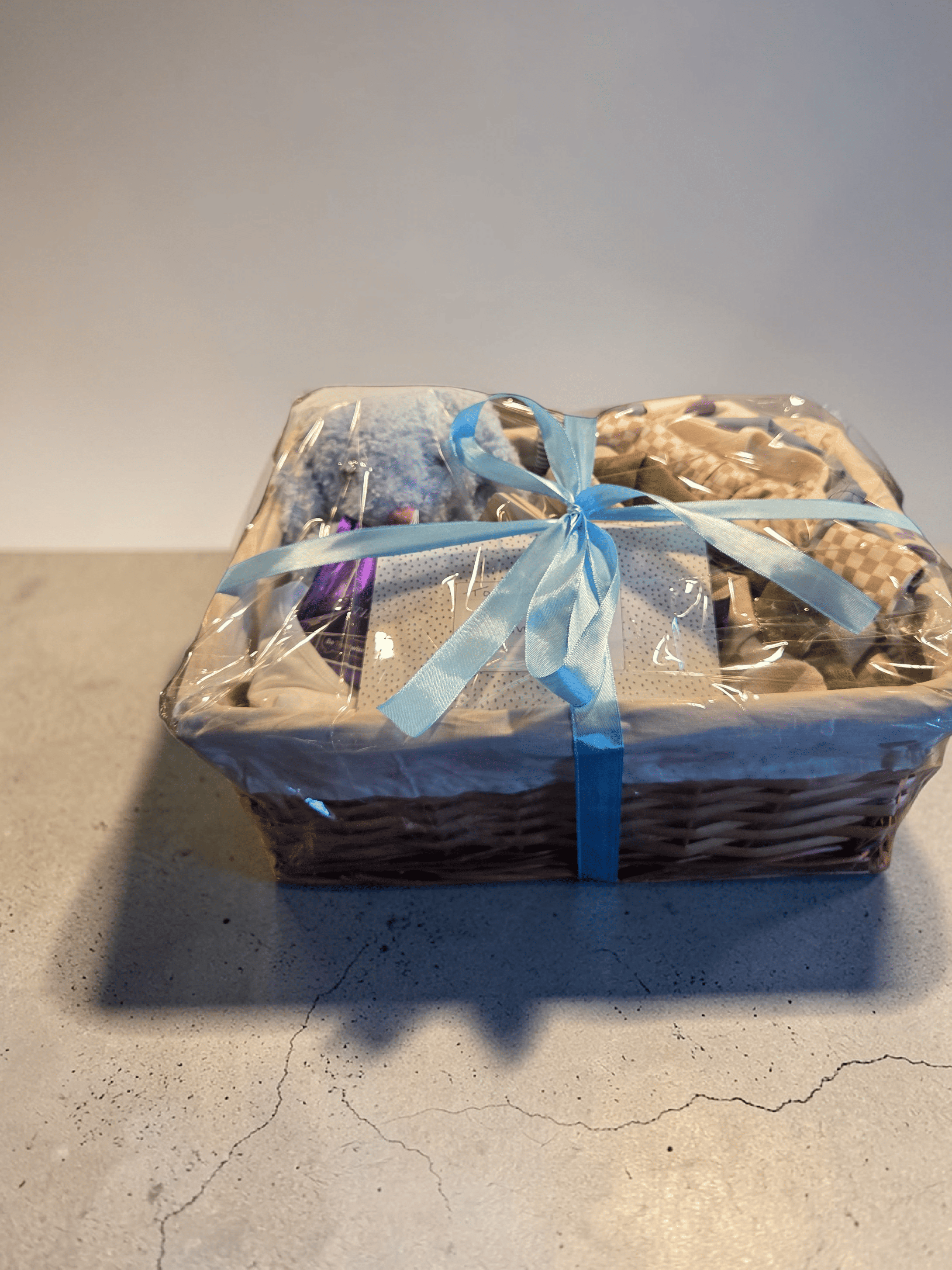 A gift basket wrapped in clear cellophane and tied with a light blue ribbon, sitting on a gray surface.