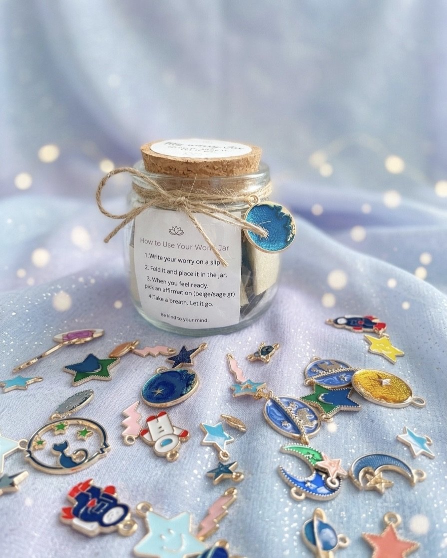 A small glass jar with a cork lid and a note attached, surrounded by assorted colorful zodiac-themed charms on a light blue fabric background with white polka dots.