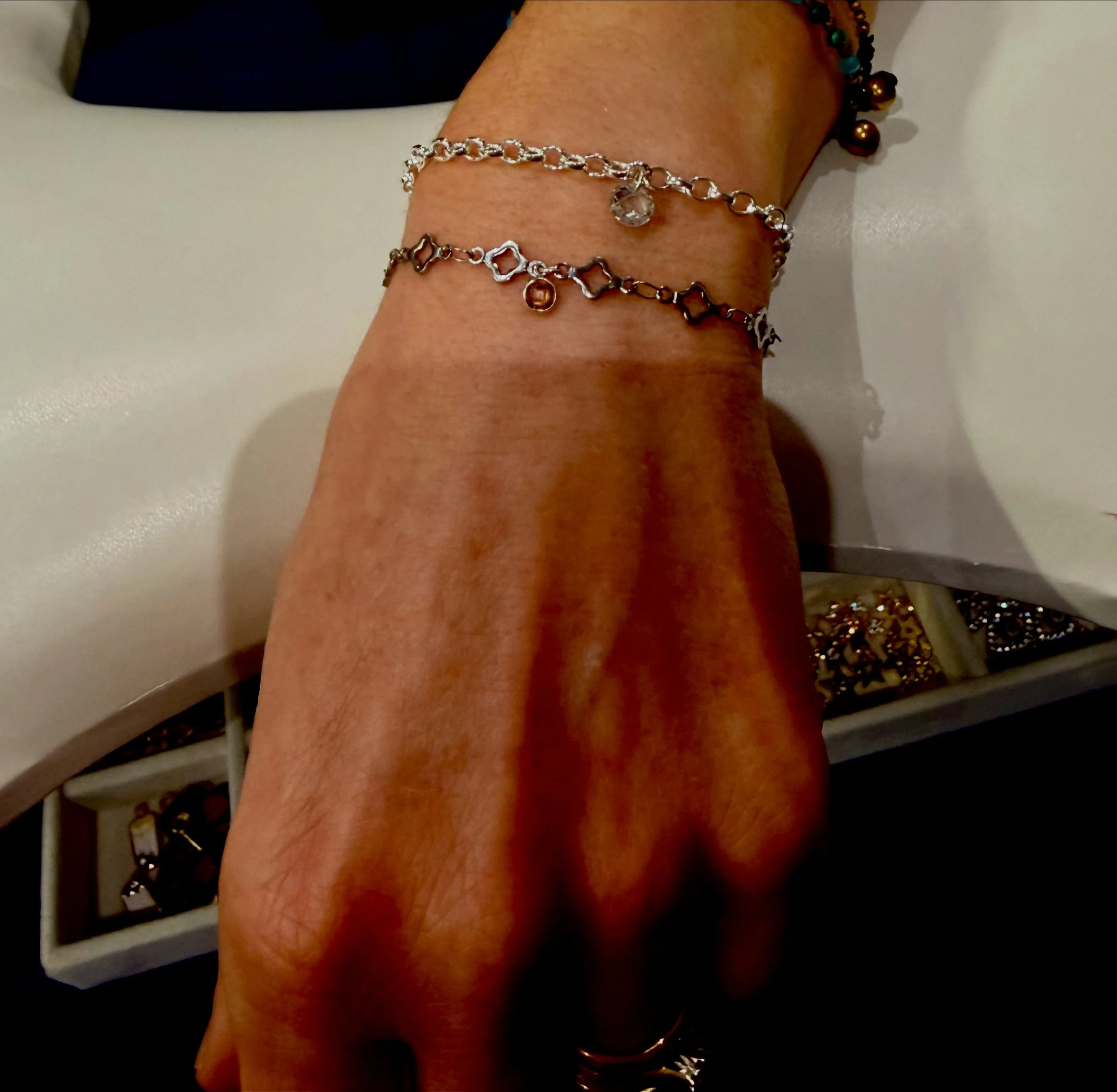 A person's arm adorned with multiple bracelets, including a silver chain bracelet with a clear central charm, a gold bracelet with a geometric pattern, and a beaded bracelet, resting on a white surface next to a container filled with various jewelry.