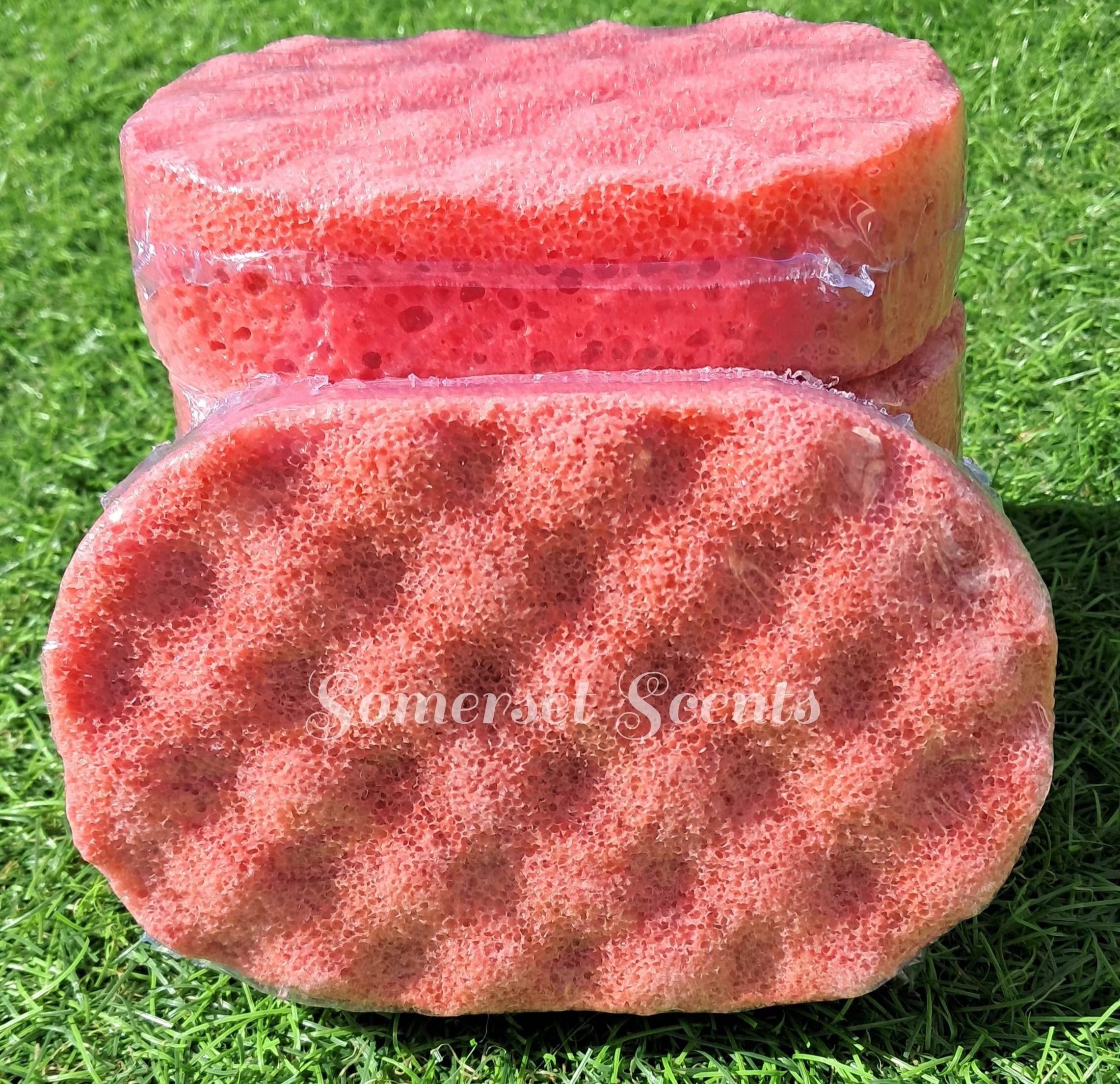 Close-up of two stacked pink and red soap bars wrapped in plastic, placed on green grass, with faint white text reading 'Somer’s Scents' visible on the soap in the foreground.