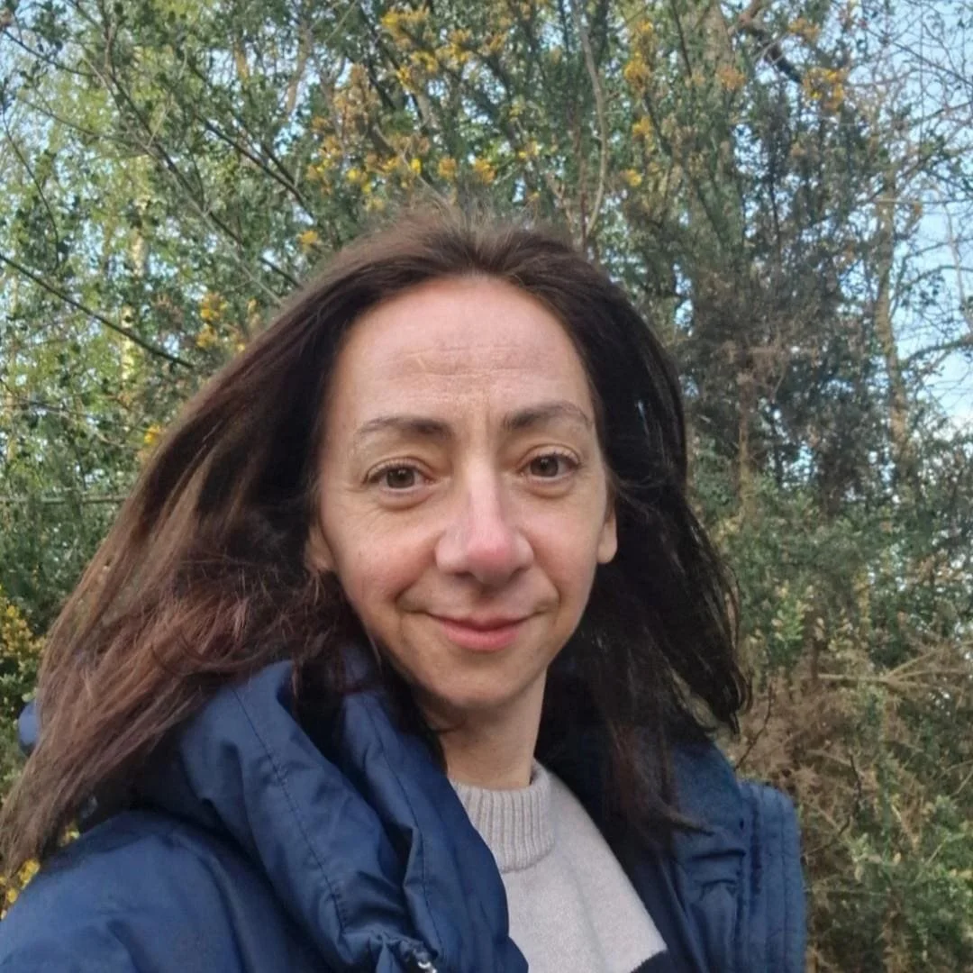 A woman with long dark hair smiling outdoors in front of trees with yellow and green leaves, wearing a gray shirt and a blue jacket.