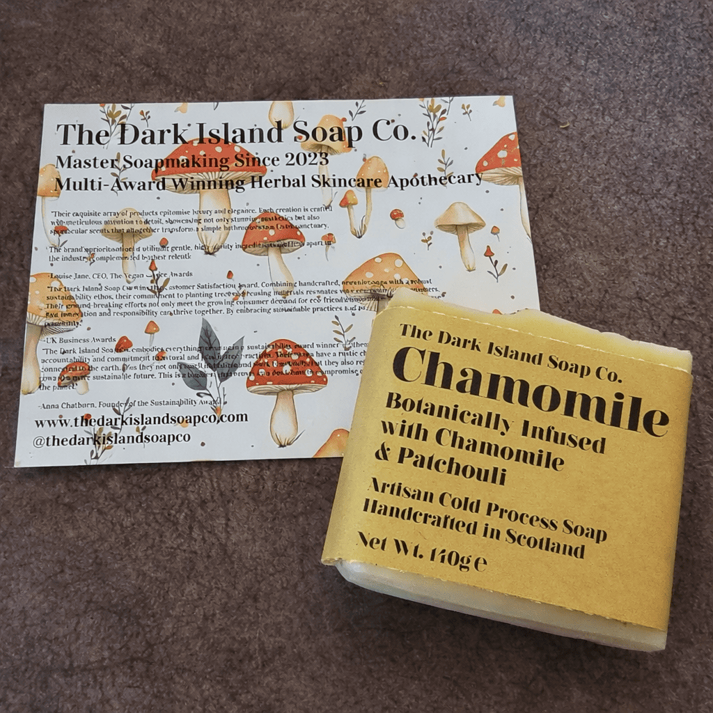 A business card and a soap packaging box from The Dark Island Soap Co. featured on a brown surface. The card has a design with mushrooms and plants, and the soap box is labeled chamomile with herbal ingredients.