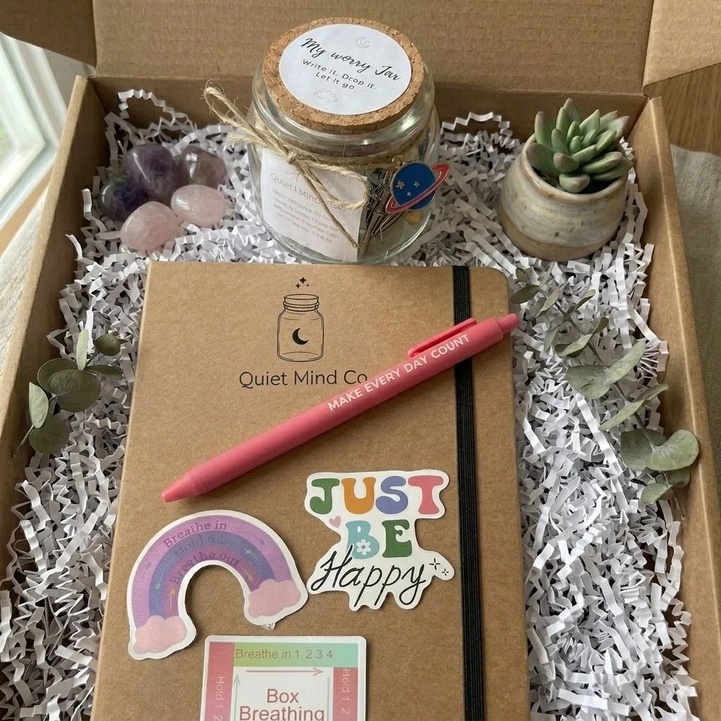 Brown gift box with white shredded paper filler containing a pink 'Make Every Day Count' pen, a 'Quiet Mind Co.' journal, a small potted succulent, a glass jar with a cork lid labeled 'My Worry Jar' with notes inside, and colorful stickers including a rainbow and the words 'Just Be Happy' and 'Breathe in 1 2 3 4'.