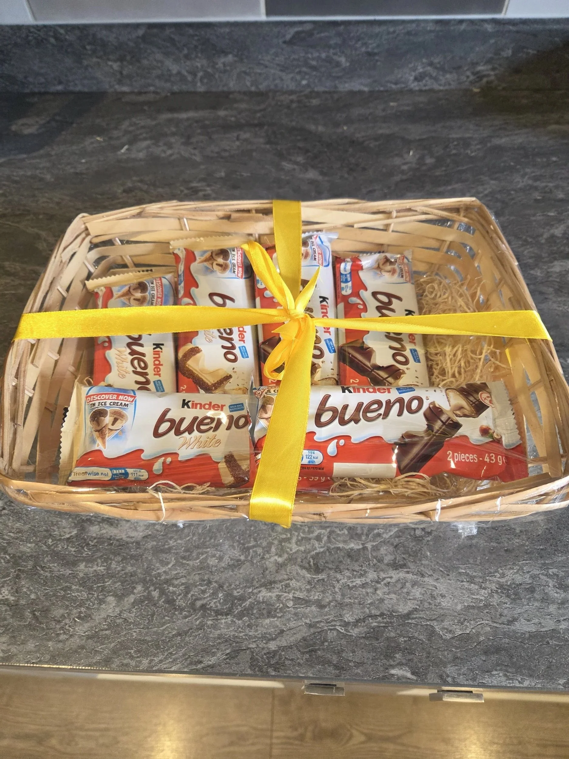 A wicker gift basket tied with a yellow ribbon, containing multiple packages of Kinder Bueno White chocolate bars, on a dark countertop.