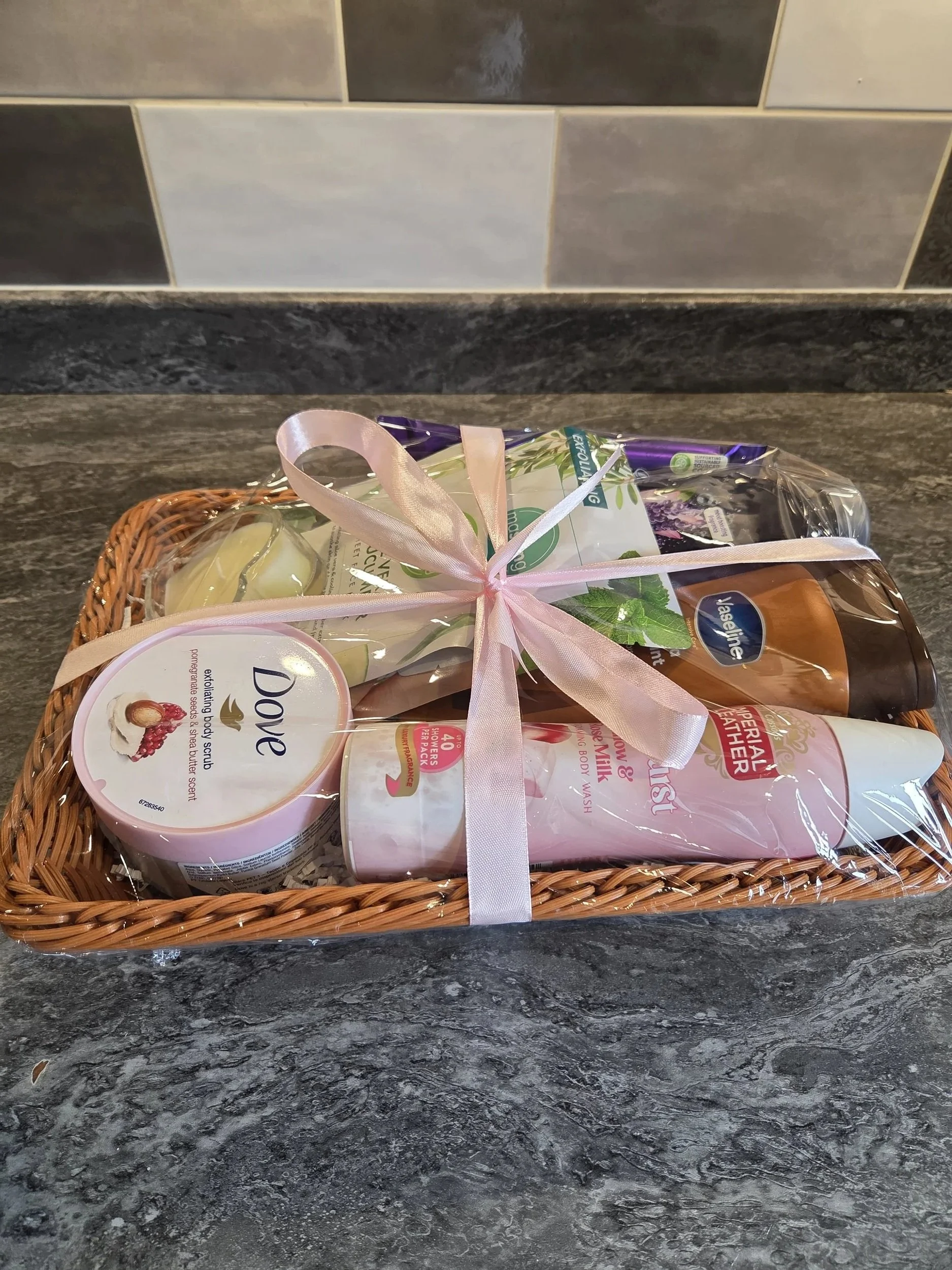 A woven basket wrapped with a pink ribbon, containing various personal care products including body scrub, lotion, and soap, placed on a dark marble countertop against a background of tiled wall.