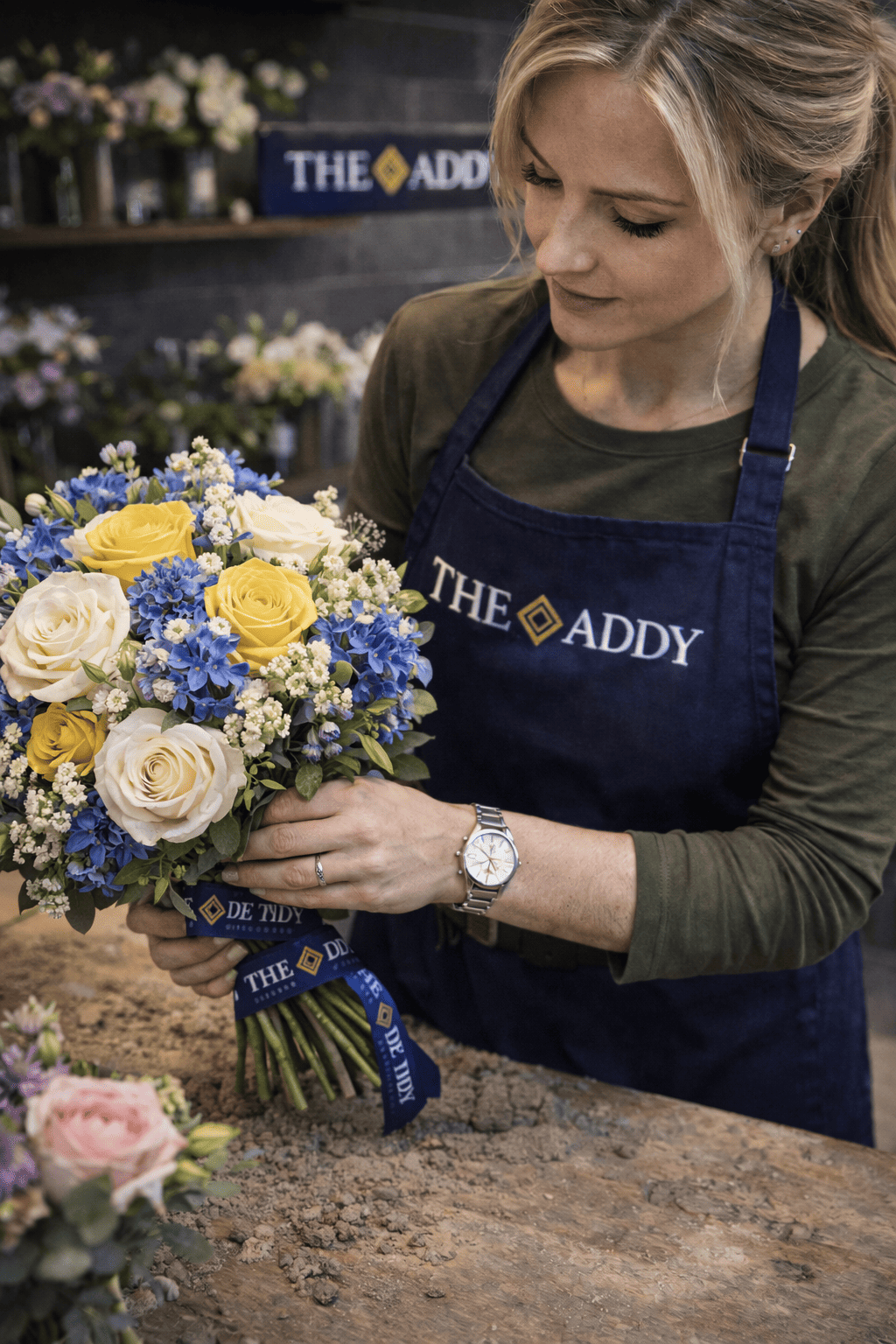 A woman in a blue apron with 'THE ADDY' logo arranging a colorful bouquet of yellow, white, and blue flowers in a flower shop.