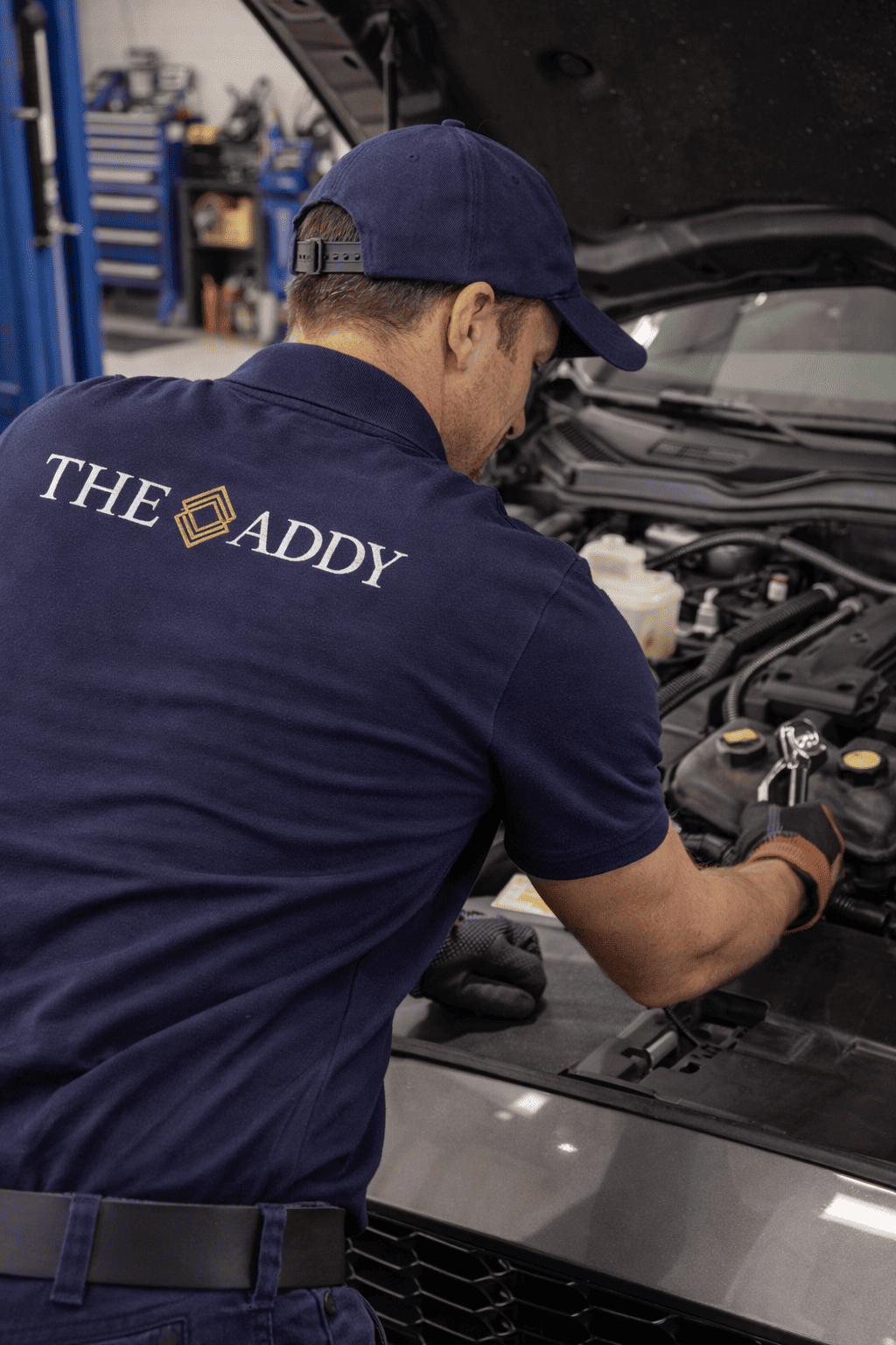 Auto mechanic working on car engine in garage, wearing a navy blue shirt with 'THE ADDY' logo on back, and navy blue cap.