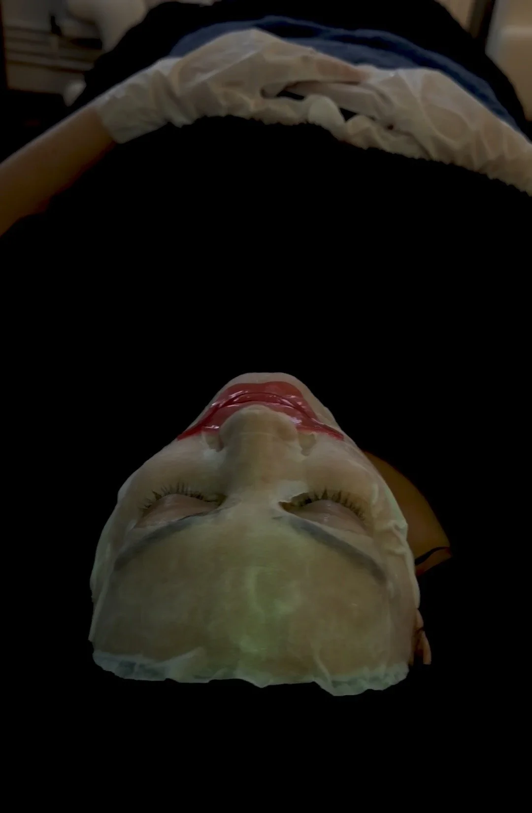 A person lying on a bed wearing a facial mask with eye and lip makeup, eyes closed.