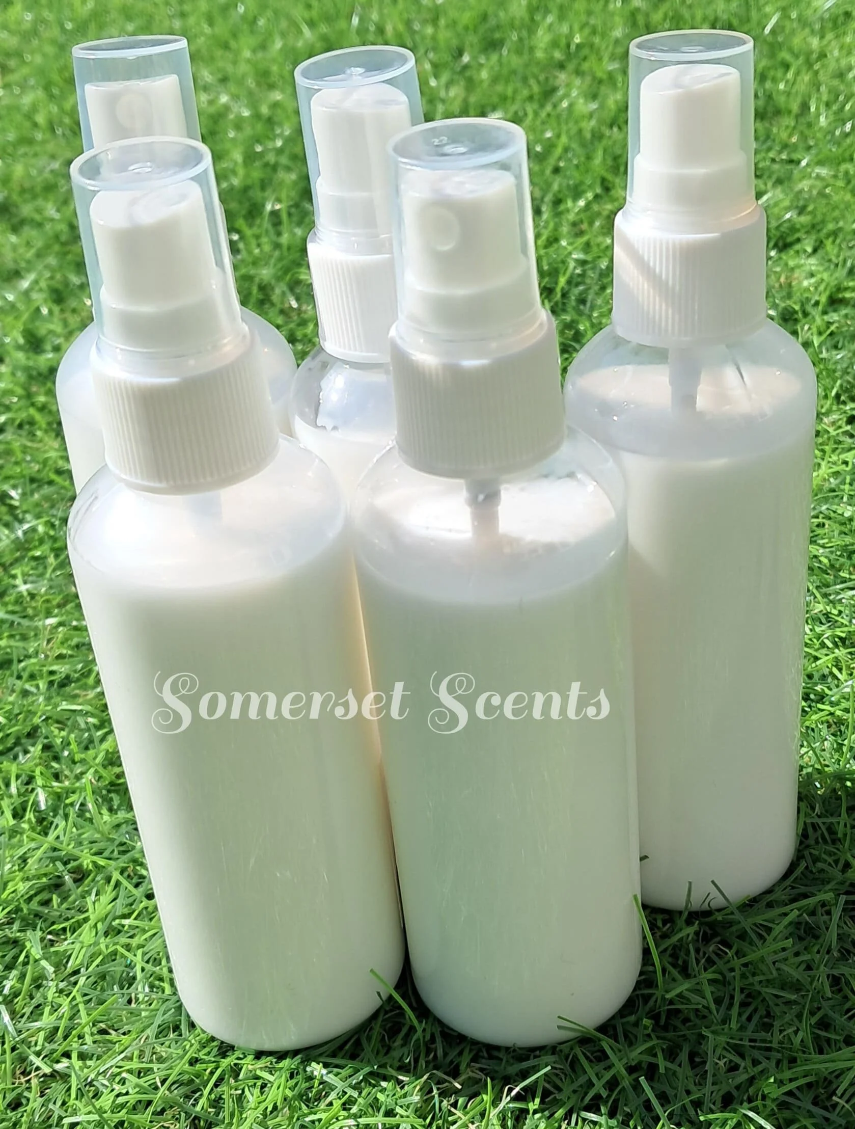 Several white spray bottles with clear caps on a green grassy surface, with a logo or text reading "Somerset Scents" overlaid at the bottom.