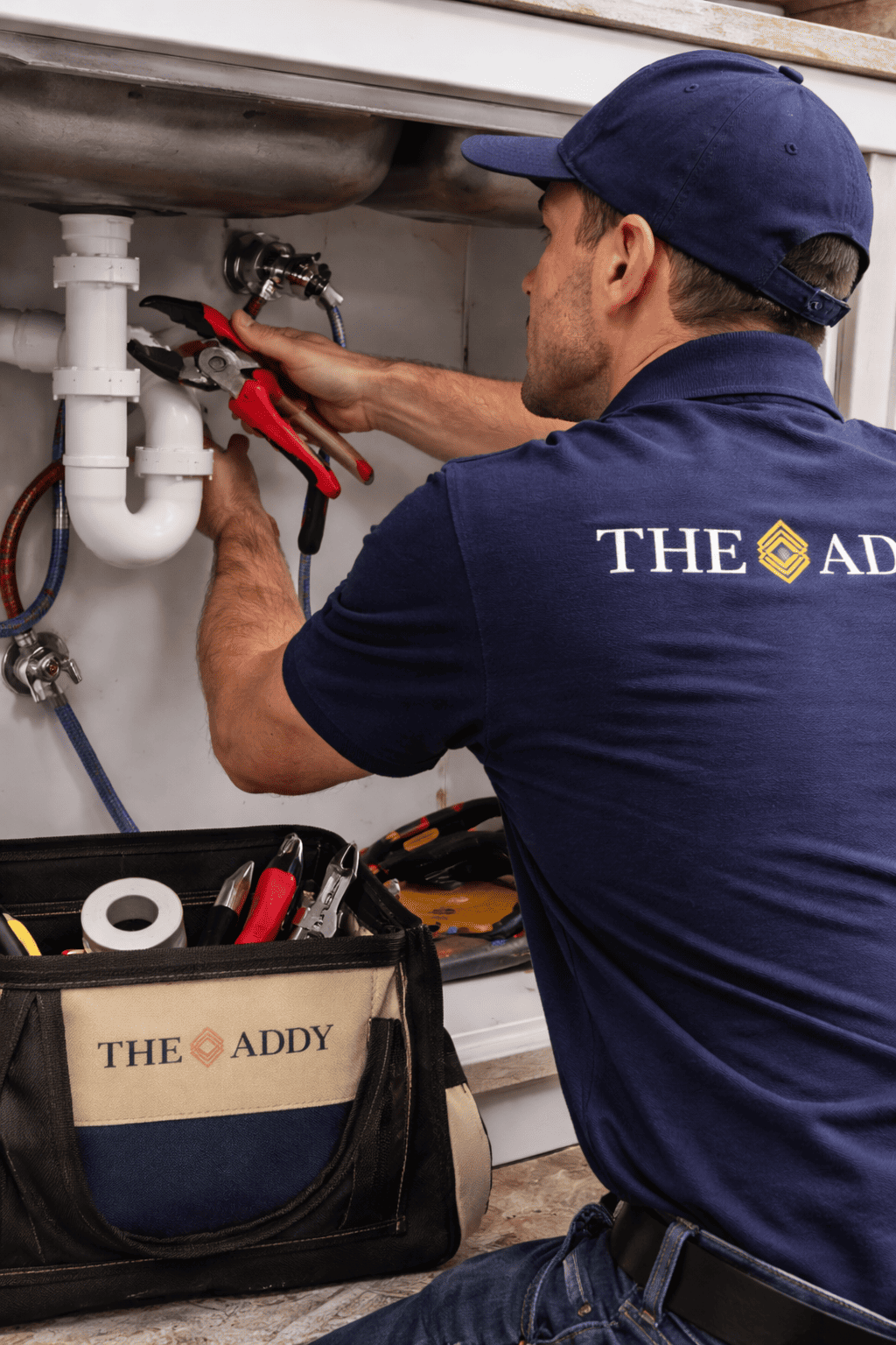 A plumber wearing a dark blue cap and uniform is working under a kitchen sink, using tools to repair plumbing pipes. A toolbox labeled 'THE ADDY' is on the floor nearby.