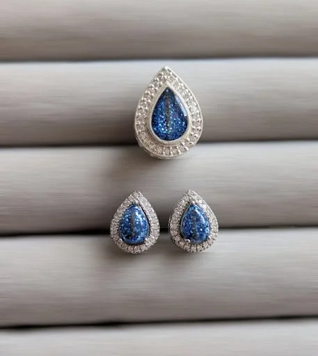 A jewelry set with a teardrop-shaped blue gemstone surrounded by small diamonds, consisting of a matching ring and earrings, displayed on a gray textured surface.