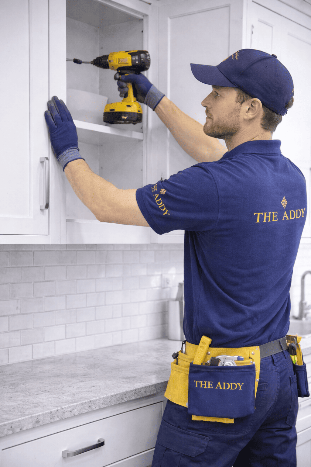 A man in a blue uniform and cap marked 'The Addy' is installing or repairing a kitchen cabinet with a cordless drill. He is wearing gloves and a tool belt.