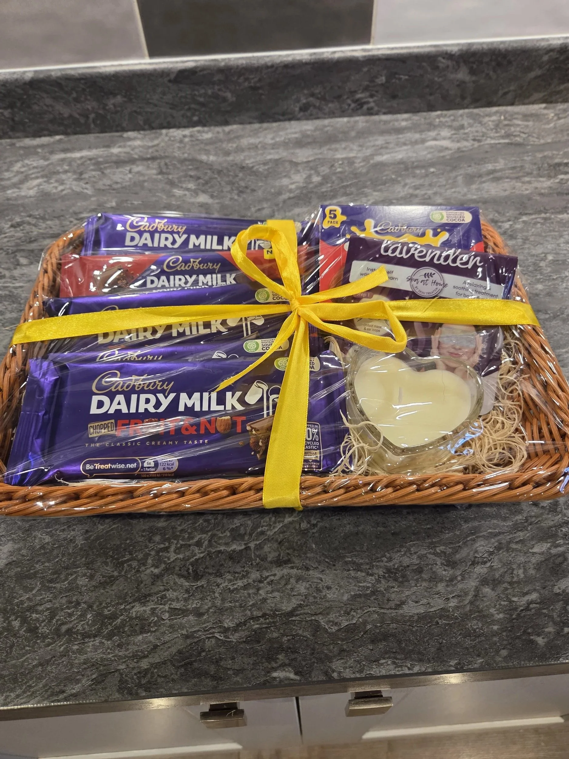 A wicker gift basket wrapped with a yellow ribbon, containing multiple packages of Cadbury Dairy Milk chocolate bars, lavender lotion, and a small candle, placed on a dark gray kitchen countertop.