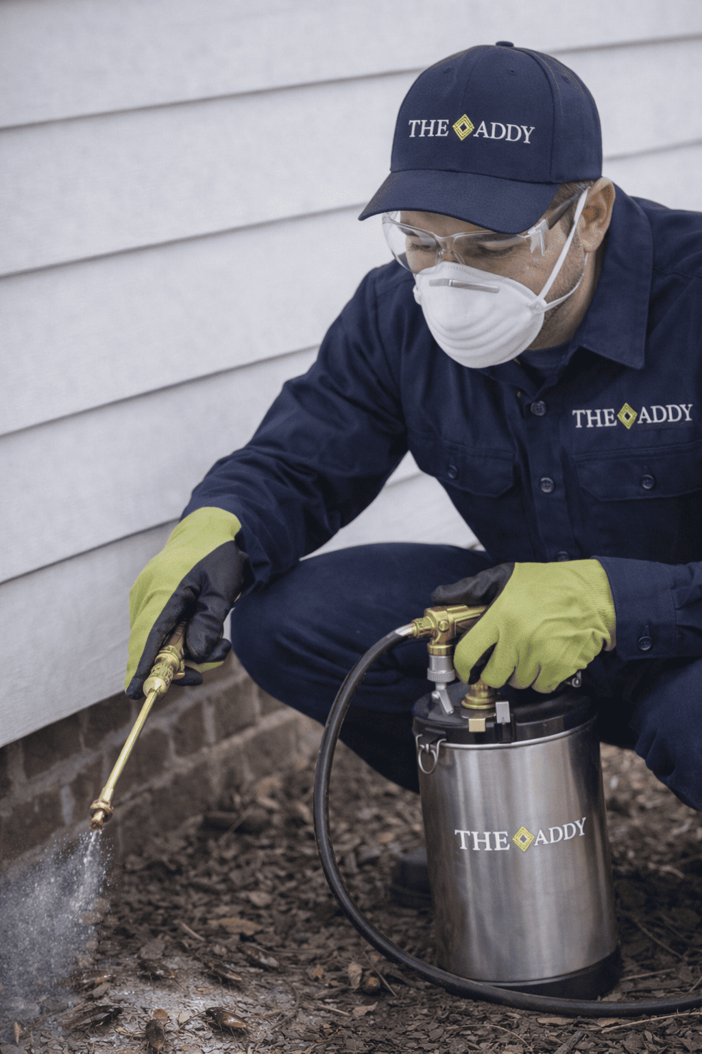 A pest control technician wearing safety gear, including gloves, goggles, face mask, and hat, is spraying pesticide near the foundation of a house.