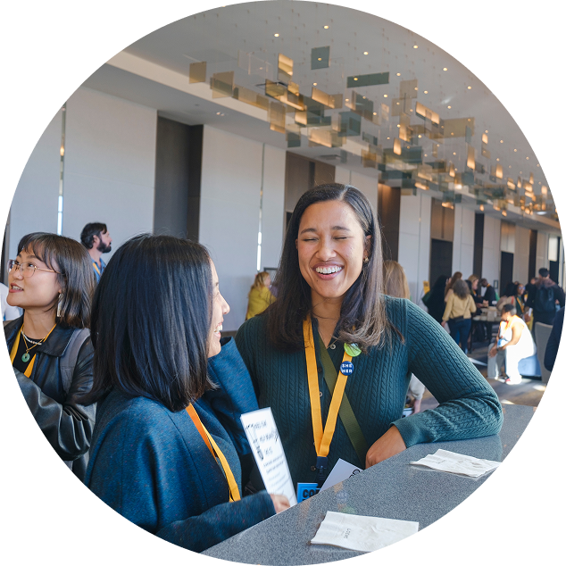 Two women smiling and talking at a conference registration desk surrounded by other attendees in a modern, well-lit venue.