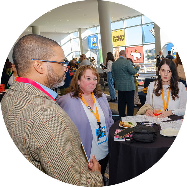 People at a conference or event in a modern venue with large windows, some are engaging in conversation near a black table with food and drinks.