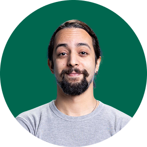 A man with long dark hair, a beard, and mustache wearing a light gray t-shirt, smiling in front of a green background.