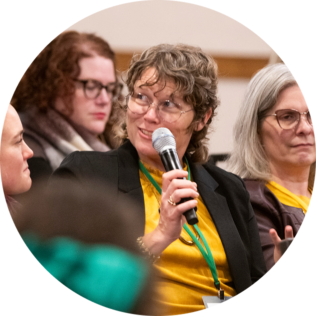 A woman with curly hair and glasses speaking into a microphone during a panel discussion or conference, with other women seated nearby.