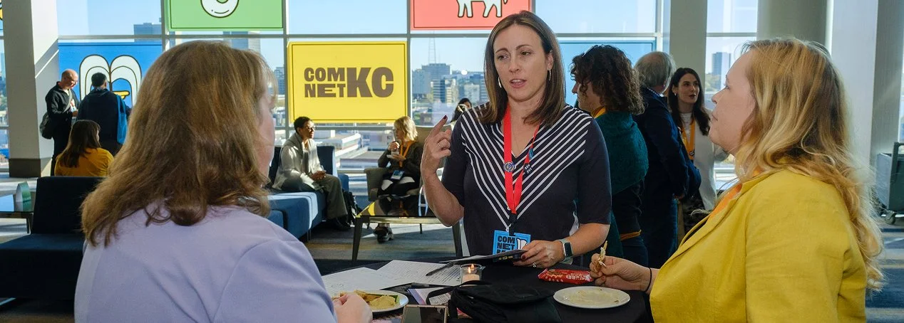 Three women engaged in conversation at a conference with a large window view of a city skyline in the background.