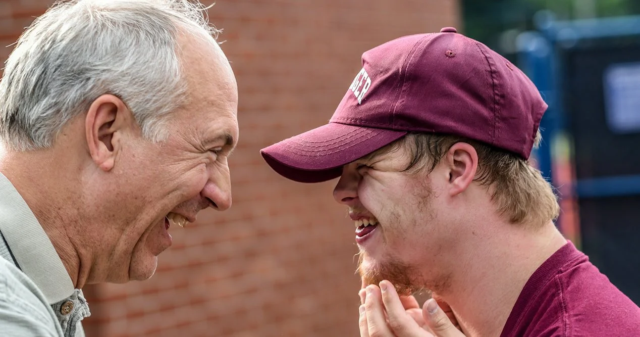 Two men smiling and laughing while touching foreheads, with a brick wall and some trees in the background.