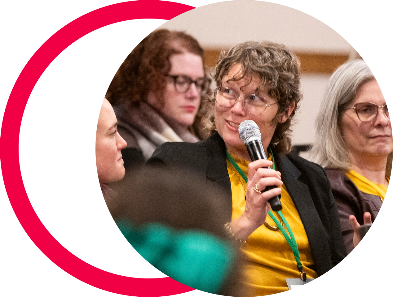 A group of women sitting in a panel discussion, one woman in the center holding a microphone and speaking, wearing glasses and a yellow shirt.